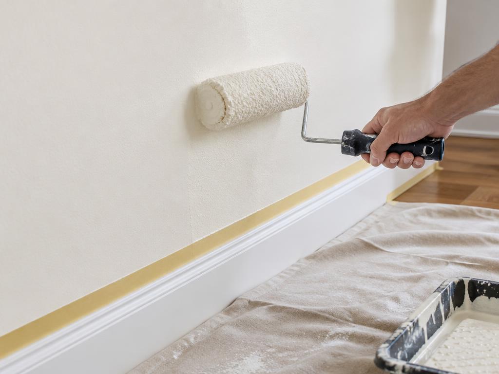 Roller applying warm white paint above taped white baseboard with drop cloth below